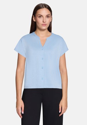 Woman wearing a light blue short-sleeve buttoned blouse with a small V-neck paired with black pants on a plain background.
