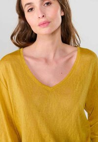 Top à manches longues en V en doux tissu texturé couleur moutarde ; coupe décontractée avec des épaules légèrement tombantes et des détails en côtes au niveau de l'ourlet.