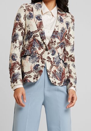 Žena nosící krémové květované sako s modrými, hnědými a fialovými akcenty, bílou košili na knoflíky a světle modré kalhoty na míru.