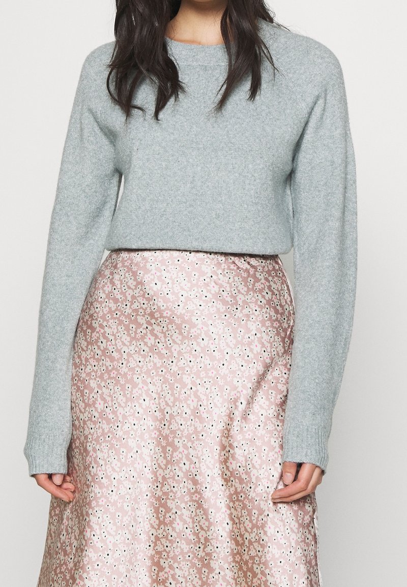 Femme portant un pull en maille gris associé à une jupe en satin à motifs floraux roses, se tenant devant un fond clair uni.