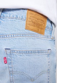 Light blue denim jeans feature a leather back patch with embossed branding, a red tab on the pocket, and contrasting stitching details.
