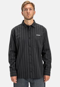 Zwart langärmige shirt met witte verticale strepen, een button-down kraag, borstzak en logo patch; gemaakt van zachte stof.