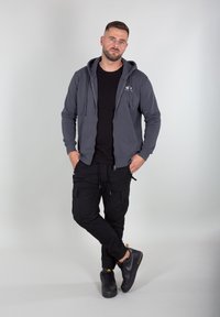 Sudadera gris con cremallera y bolsillo frontal, llevada sobre una camiseta negra y combinada con pantalones cargo negros y zapatillas negras con acentos naranjas.