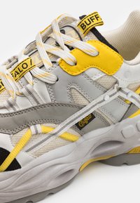 Világosszürke és sárga sneaker, vegyes anyagú felsőrésszel, velúr és hálós részletekkel, vastag középtalppal és réteges dizájnelemekkel.