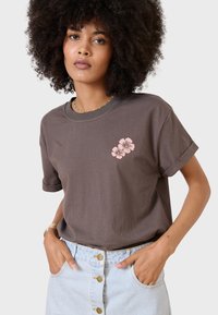 Camiseta de algodón marrón con mangas enrolladas y un diseño floral rosa en el pecho, combinada con una falda de mezclilla azul claro con cierre de botones.