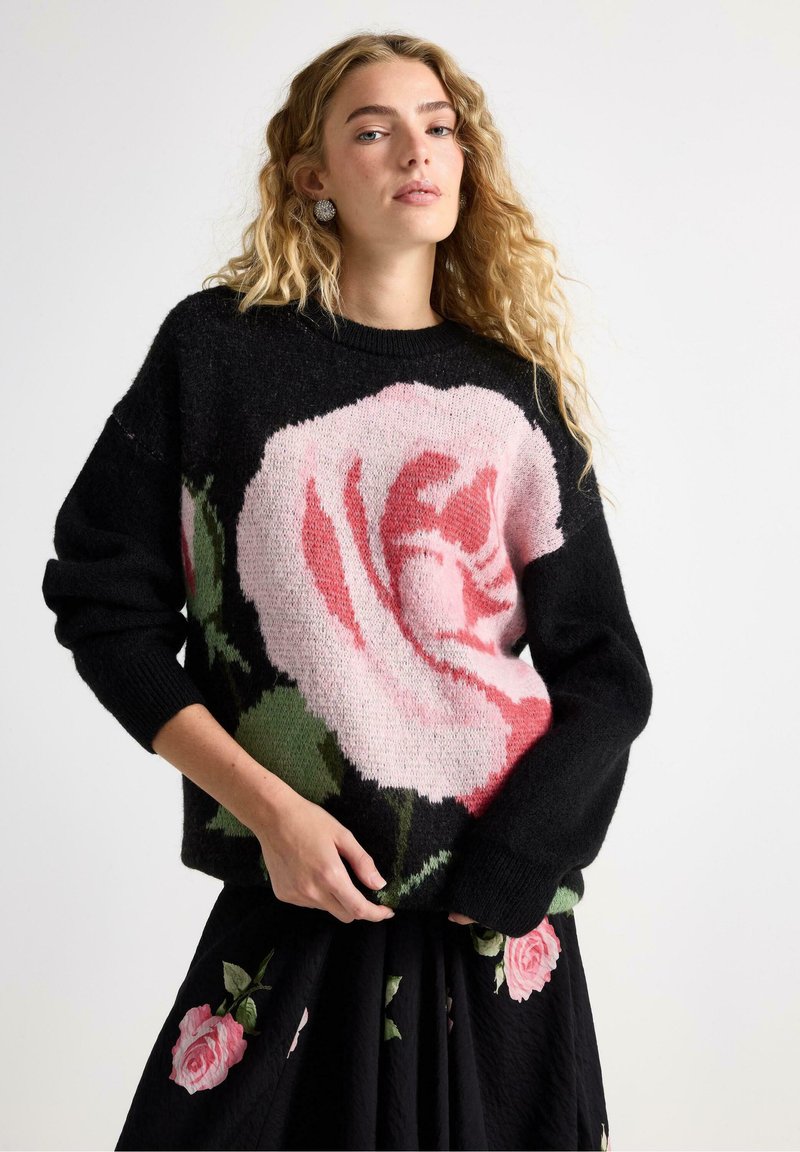 Černý svetr s velkým designem růže v odstínech růžové a zelené. Tkanina vypadá měkce, má uvolněný střih a žebrované manžety.
