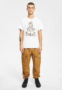 Młody mężczyzna stoi, ubrany w biały t-shirt z beżowo-czarną grafiką i napisem „Dakar”, beżowe spodnie typu cargo oraz czarne buty, na jednolitym tle.