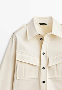 Chemise beige clair avec un design structuré, dotée d'un col pointu, de deux poches poitrine et de boutons noirs. Tissu texturé avec une finition lisse.