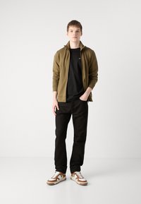 Olivengrønn zip-up jakke, svart t-skjorte, svarte jeans og brune og hvite sneakers; uformell antrekk med en avslappet passform.