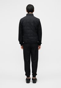 Μαύρο καπιτονέ γιλέκο με ψηλό γιακά, συνδυασμένο με μαύρα τζόγκερ με στενά τελειώματα στα μανσέτα. Ομαλό υλικό με υφή στα τζόγκερ.