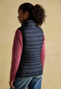 Melnā polsterētā jaka ar horizontālām šuvēm, valkāta virs rozā džempera. Ievērojama apkakle un gluda tekstūra, ar etiķeti aizmugurē.