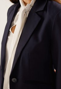 Blazer azul marino con hombros estructurados y un solo botón negro. Pañuelo blanco superpuesto en el cuello que cae con elegancia. Textura de tela lisa.