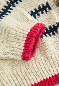 Gebreide trui met een crèmekleurige stof en een textuurpatroon, rode geribbelde mouwen en donkerblauwe accenten in een geometrisch ontwerp.