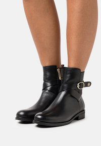 Botas de cuero negro hasta el tobillo con cremallera lateral, que presentan un detalle de hebilla y una textura suave. Diseño de tacón bajo y punta redonda.
