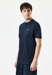 Marineblå kortærmet t-shirt lavet af blødt stof, med et lille firkantet logo på brystet, afslappet pasform og rund halsudskæring.
