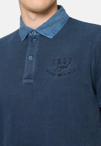 Tengerészkék pólóing texturált anyagból, kontrasztos kék gallérral, három gombbal, és hímzett "1967" részlettel a mellrészen.