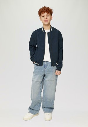 Marine bomberjack met witte gestreepte accenten, over een wit shirt. Lichtblauwe baggy jeans en witte sneakers maken de outfit compleet.