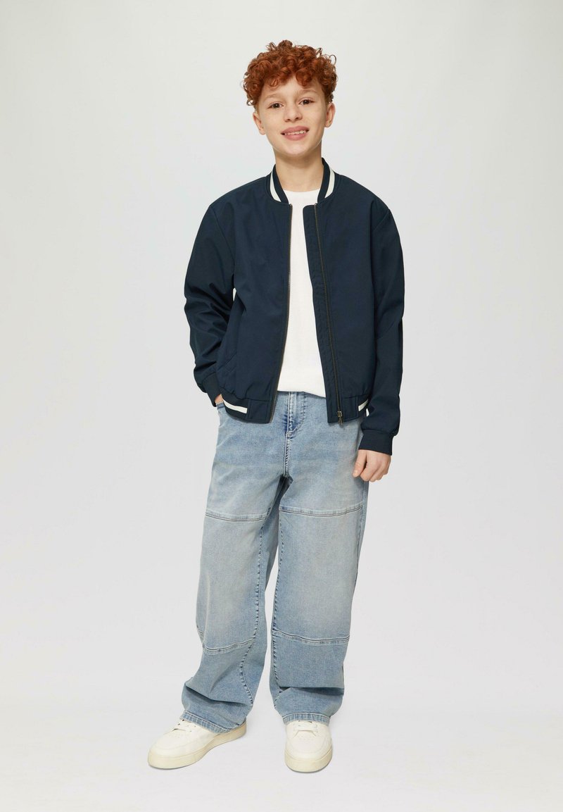 Marineblauer Bomberjacke mit weißen gestreiften Akzenten über einem weißen Hemd. Hellblaue, weite Jeans und weiße Sneakers runden das Outfit ab.