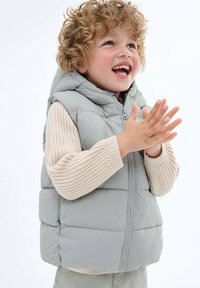 Jasnoszary kamizelka puchowa z kapturem, z suwakiem z przodu i poziomym przeszyciem, noszona na beżowym swetrze z prążkami.