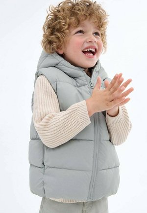 Vest puf gri deschis cu glugă, având un fermoar frontal și cusături orizontale, purtat peste un pulover bej cu model în relief.