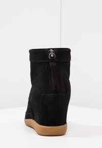 Svart mocka kilklackankelhög med en bakre dragkedja, rundad övre kant och kontrasterande brun gummisula. Texturerat material med sömnadsdetaljer.