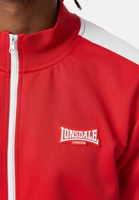 Sarkans sporta jakas ar augstu apkakli, baltas akcenti uz pleciem, priekšējais rāvējslēdzis un izteikta Lonsdale London logo uz krūtīm.