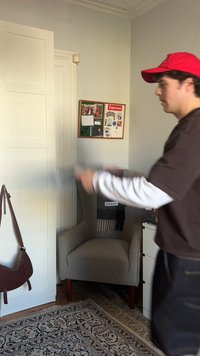 Ein junger Mann mit einer roten Mütze und geschichteten braunen und weißen Hemden steht neben einem grauen Sessel, einem Schrank und einer Pinnwand mit Aufklebern.