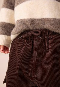 Bruna kordfläskbyxor med en strukturerad ribbdesign och midjeresår med snörning, tillsammans med en randig stickad tröja i gräddvit och grå.