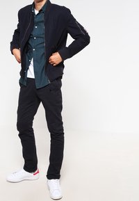 Marineblauwe bomberjack met ritssluiting, groen geruit overhemd, wit T-shirt eronder, zwarte broek en witte sneakers met rode accenten.