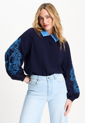 Vrouw met lang blond haar draagt een marineblouse met felblauwe bloemige borduursels op mouwen en kraag, gecombineerd met lichtblauwe spijkerbroek.
