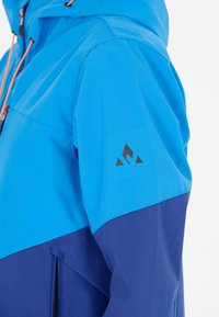 Blå och marin färgblockerad jacka med dragkedja fram, snörning i huvan och svart logotyp på ärmen. Slät, lättviktsmaterial.