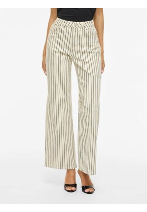 Žena stojící na bílém pozadí, oblečená v béžových širokých kalhotách s vysokým pasem a tenkými svislými černými pruhy, a v černých sandálech s otevřenou špičkou.