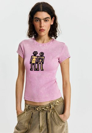 Vrouw met krullend donker haar, gekleed in een roze nauwsluitend T-shirt met abstracte figuren en een bruine cargobroek met een gouden kettingriem.