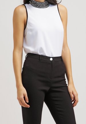 Femme portant un haut blanc sans manches avec un col orné de perles noires et un pantalon noir taille haute, debout devant un fond uni.