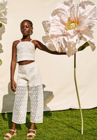 Top corto in pizzo bianco e pantaloni a gamba larga con motivi geometrici, abbinati a sandali beige. La modella tiene un grande fiore finto.