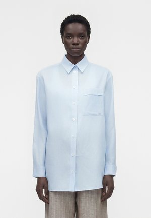 Άτομο στέκεται μπροστά φοράει ανοιχτό μπλε πουκάμισο με κουμπιά και τσέπη στο στήθος και μπεζ παντελόνι με ρίγες.