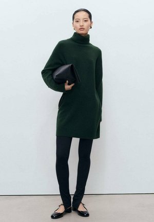 Vrouw in een donkergroene coltrui jurk, zwarte legging, zwarte schoenen met lage hakken, die een grote zwarte clutchtas tegen een lege muur houdt.