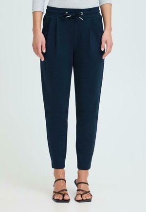 Femme portant un pantalon fuselé bleu marine avec taille à cordon et plis, associé à des sandales noires à lanières et bouts ouverts, se tenant devant un fond blanc.