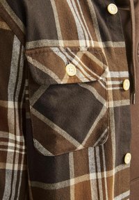 Detailní záběr kapsy hnědé kárované košile s krémovým knoflíkem a odpovídajícími krémovými knoflíky na přední straně košile.