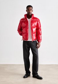 Red puffer jacket with a shiny texture, zip closure, and a high collar, paired with a light shirt and dark jeans, standing on a neutral background.
