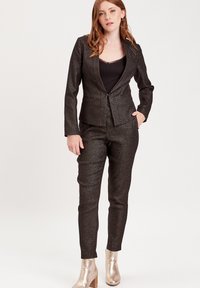 Black, textured suit with fitted blazer and slim trousers. Shiny pattern, layered over a black top. Metallic ankle boots with block heels.