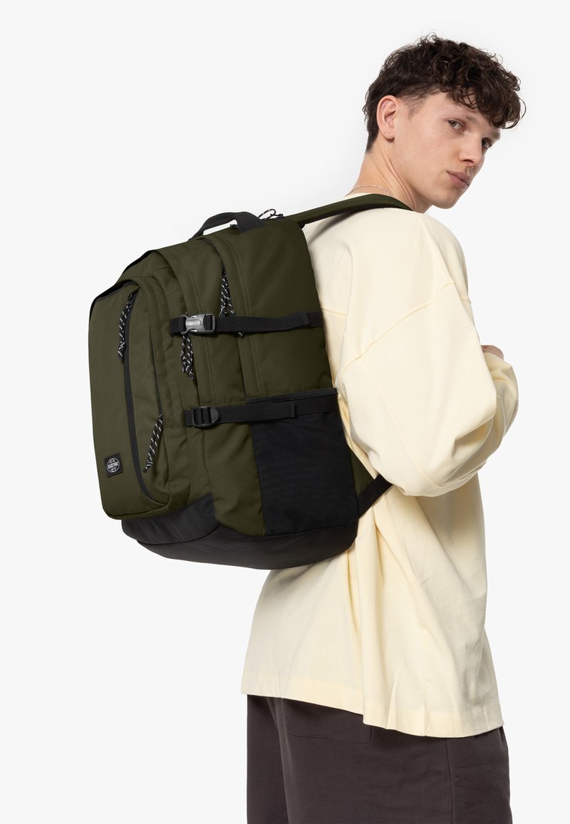 Olivgrön ryggsäck med svarta accenter, flera förvaringsfack, vadderade bärremmar och texturerad detaljering på nedre delen.
