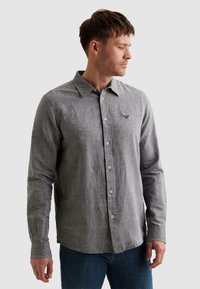 Homme portant une chemise grise à manches longues boutonnée et un jean bleu, regardant vers sa droite sur un fond clair uni.