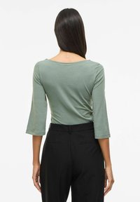 Grünes, tailliertes Oberteil mit dreiviertel Ärmeln aus leichtem Stoff. Kombiniert mit hoch taillierter schwarzer Hose. Minimale Steppnähte.