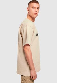 Béžové bavlněné tričko s krátkým rukávem, volný střih, s černým textem "2Y" na hrudi. Hladká textura, jednoduchý design.