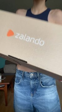 Zalando kartondoboz, elöl logóval; világosbarna szín; egy személy előtt tartva, aki kék felsőt és farmer rövidnadrágot visel.