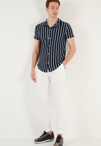 Camicetta a righe navy a maniche corte con strisce bianche, chiusura con bottoni, vestibilità comoda, abbinata a pantaloni bianchi e sneakers scure.
