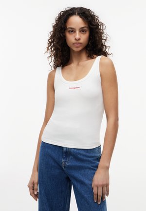 Kobieta z kręconymi włosami, ubrana w biały prążkowany top na ramiączkach z czerwonym logo "tommy jeans" i niebieskie dżinsy, stojąca na białym tle.