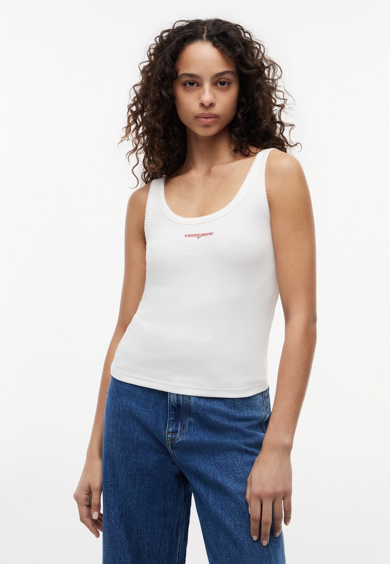 Femme aux cheveux bouclés portant un débardeur blanc côtelé avec le logo rouge "tommy jeans" et un jean bleu, debout devant un fond blanc.