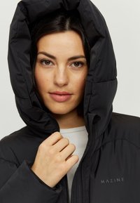 Black puffer jacket with a large hood, featuring a zip front and sleek design. Material appears soft with a smooth texture. Logo visible on chest.